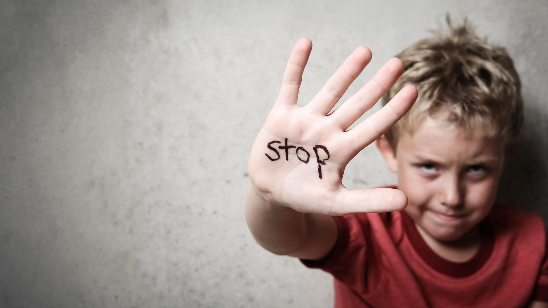 Young boy sitting against a wall, upset and holding up a hand that has the word “STOP” written inside his palm