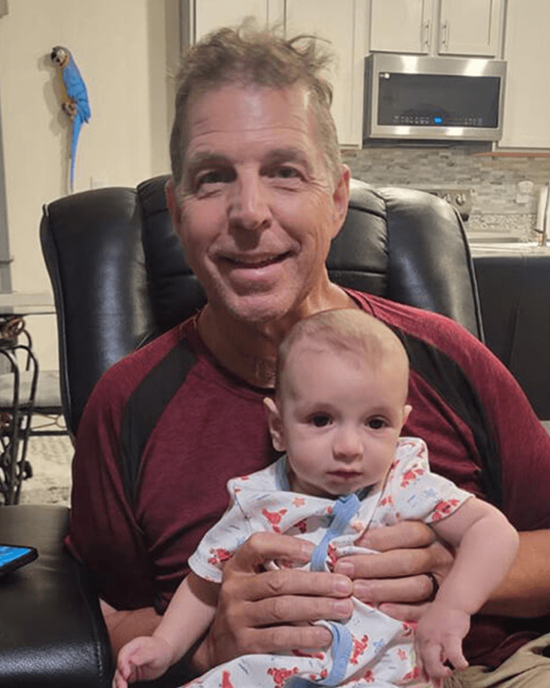 James Roger smiling and sitting in a recliner chair in a living room holding an infant on his lap